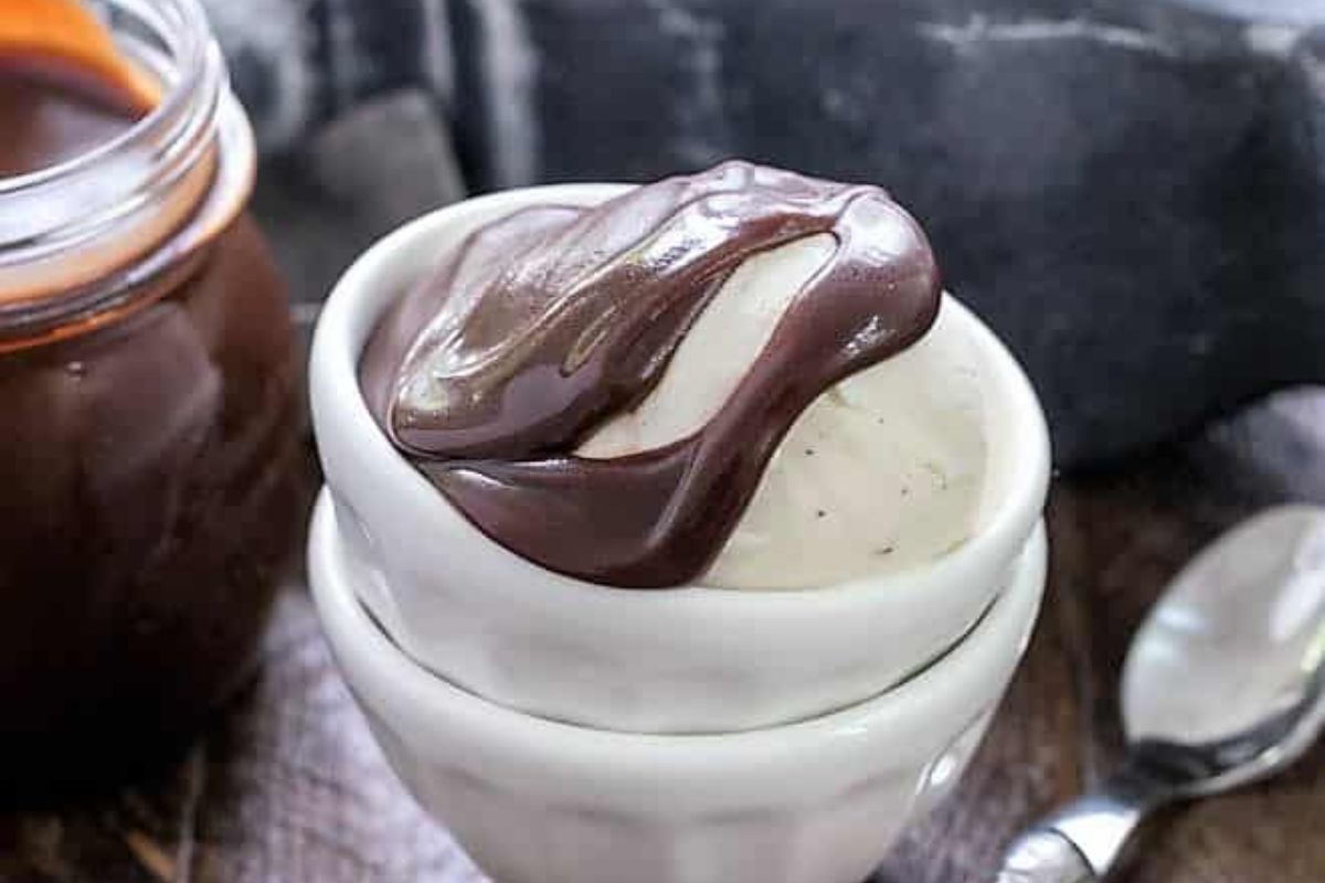 A bowl of vanilla ice cream topped with chocolate sauce, next to a jar of sauce and a spoon on a wooden surface.