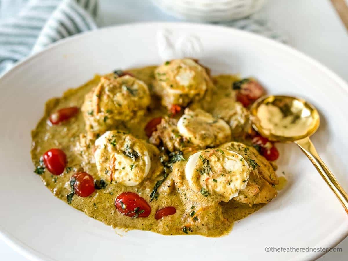 A white plate with chicken breasts in a creamy herb sauce, garnished with cherry tomatoes, served with a gold spoon.