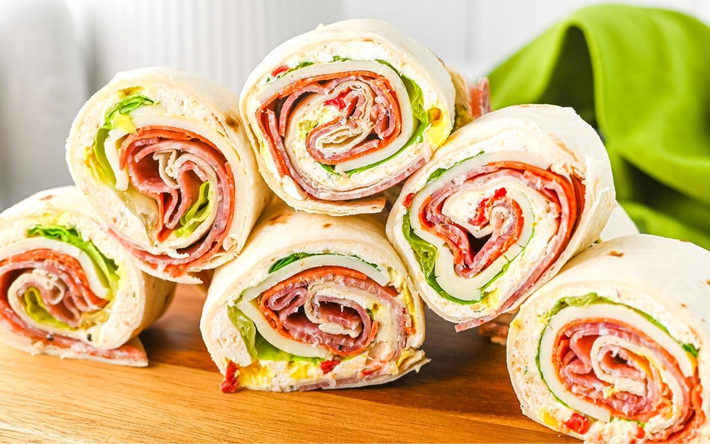 Six pinwheel sandwiches with deli meat, cheese, lettuce, and tortillas are stacked on a wooden board with a green cloth in the background.