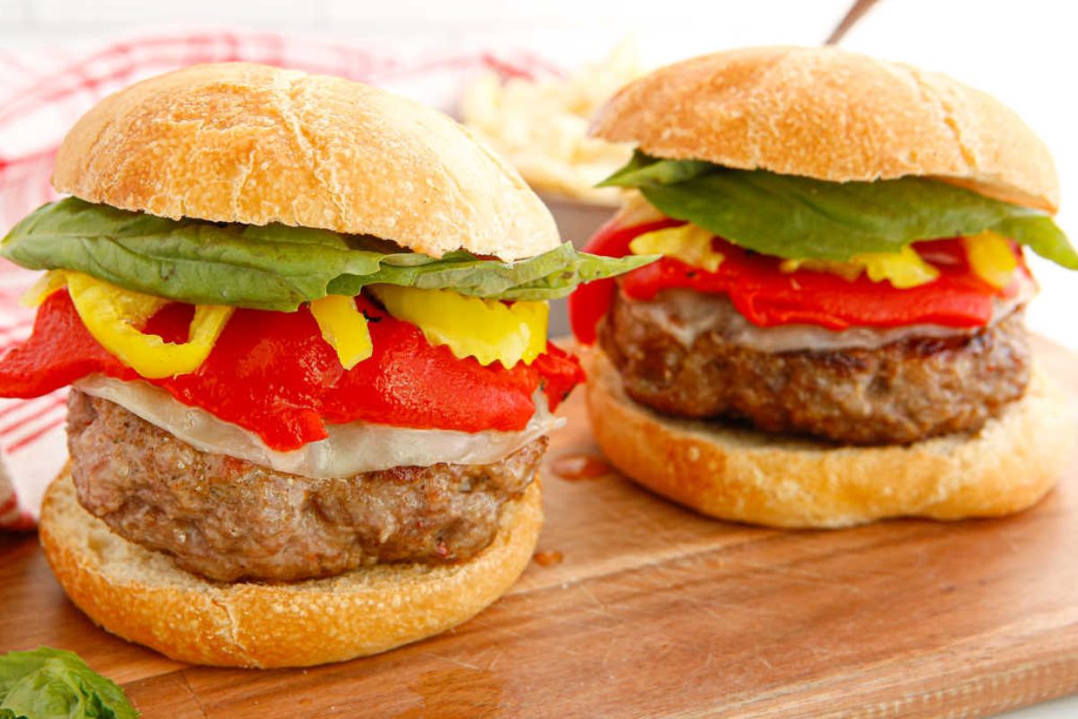 Two hamburgers with beef patties, lettuce, sliced yellow peppers, roasted red peppers, and cheese on sandwich buns, served on a wooden board.