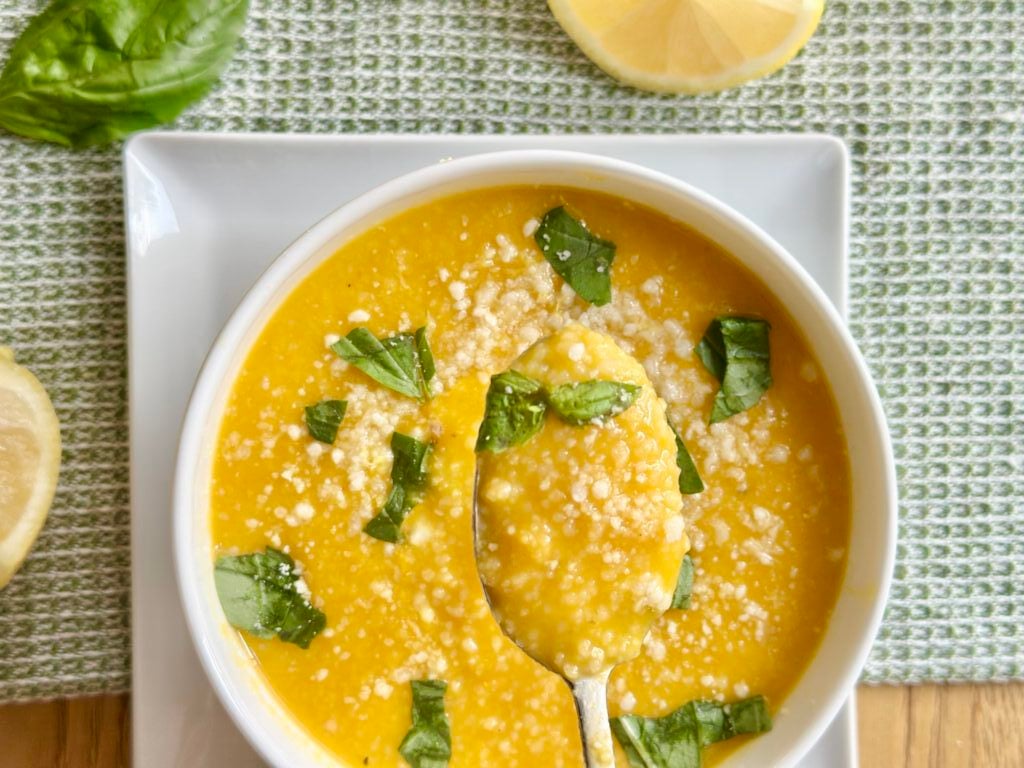 Bowl of yellow soup topped with grated cheese and fresh basil, with a spoon, on a white plate and green textured placemat. Lemon slices and basil leaves nearby.