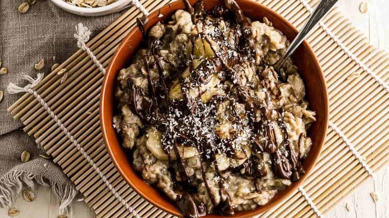 A bowl of oatmeal topped with shredded coconut and drizzled with chocolate sauce, placed on a bamboo mat with a spoon inside.