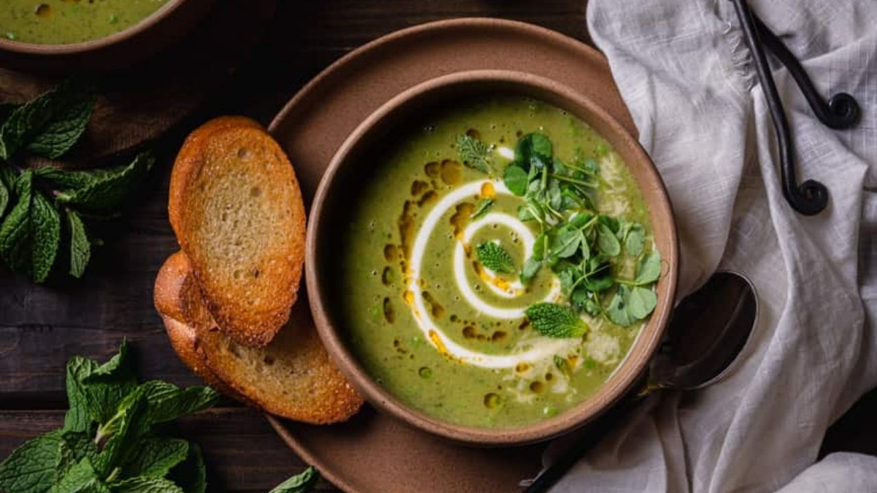 A bowl of green soup garnished with cream, herbs, and oil, served with two slices of toasted bread on a brown plate, next to a napkin, spoon, and fresh mint leaves.