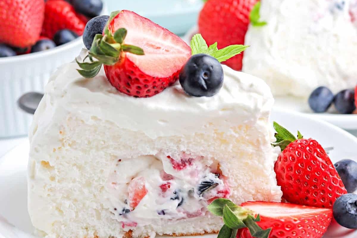 A slice of white cake with whipped cream, topped and filled with fresh strawberries and blueberries, served on a white plate.