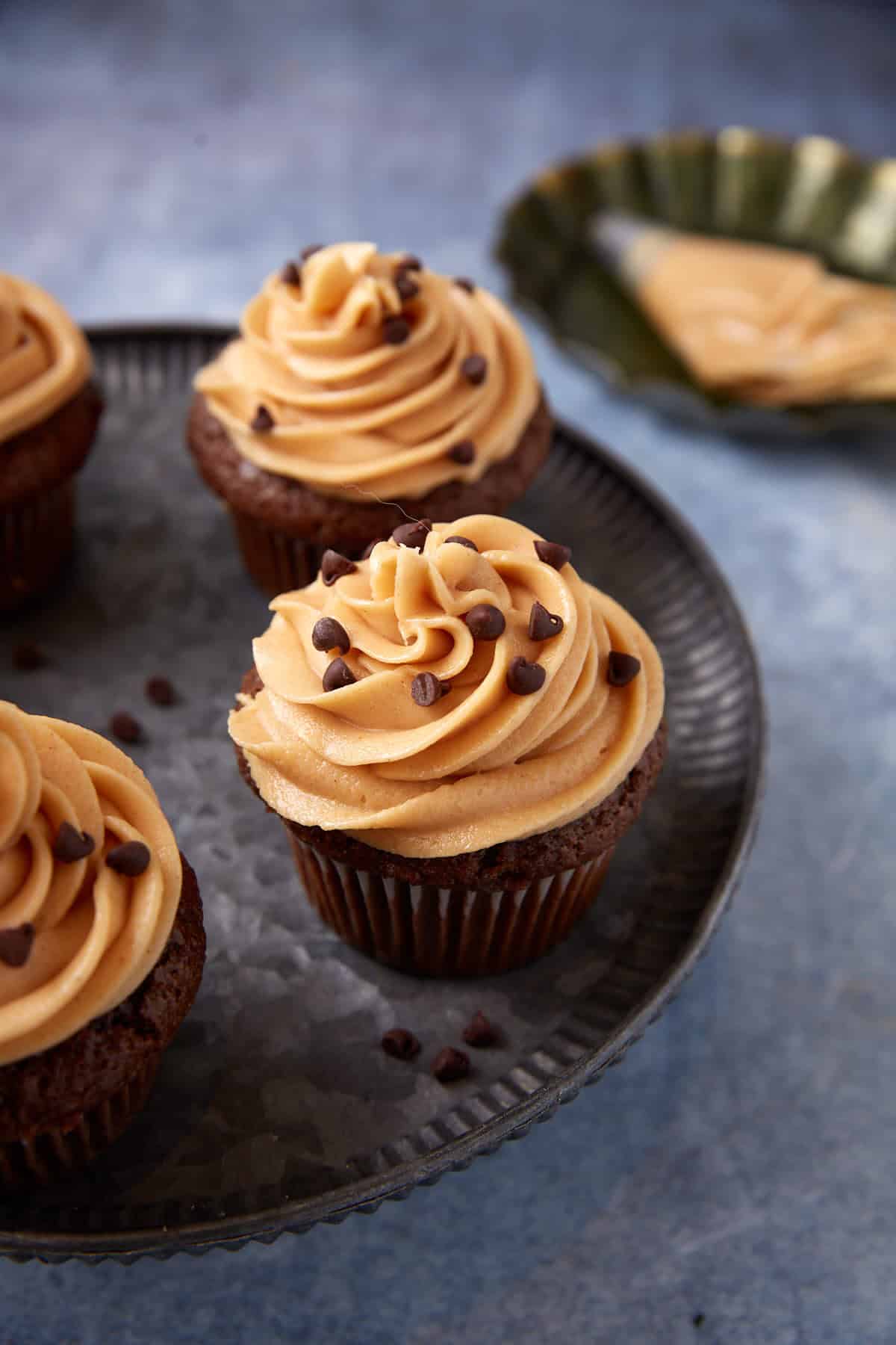 Three chocolate cupcakes topped with swirled peanut butter frosting and chocolate chips on a metal tray, perfect for fans of irresistible peanut butter recipes. Frosting and utensils are scattered in the background.
