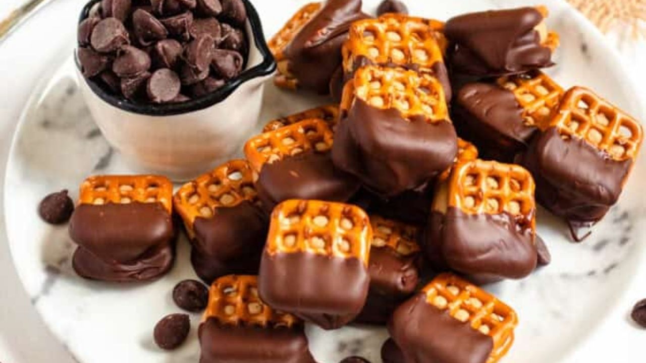 A plate of pretzel sandwiches partially dipped in chocolate, next to a small cup filled with chocolate chips.