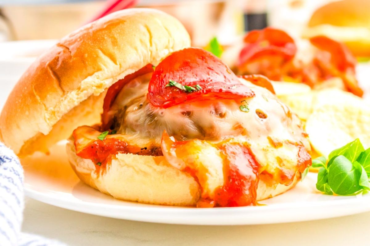A hamburger topped with melted cheese, tomato sauce, and pepperoni on a bun, served with chips and a garnish on a white plate.
