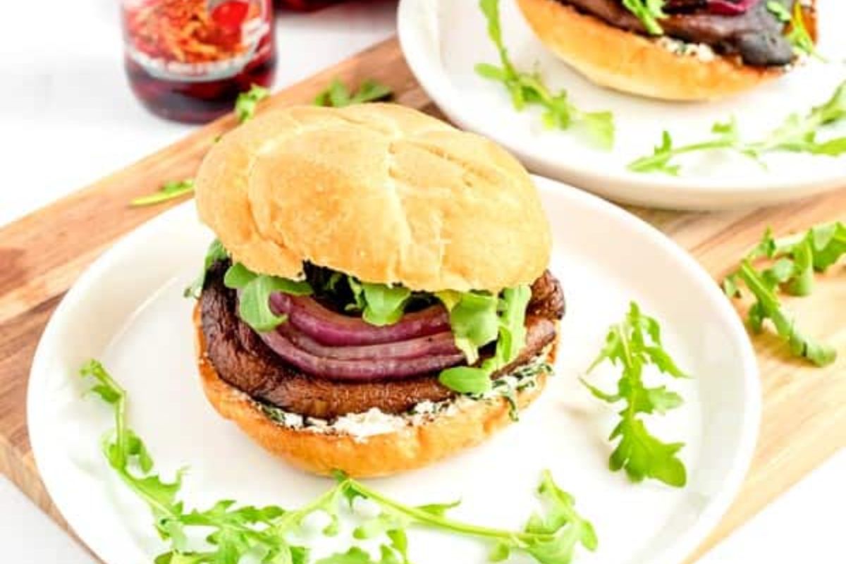A sandwich with a large grilled portobello mushroom, red onion, arugula, and a creamy spread on a bun, served on a white plate with arugula garnish.
