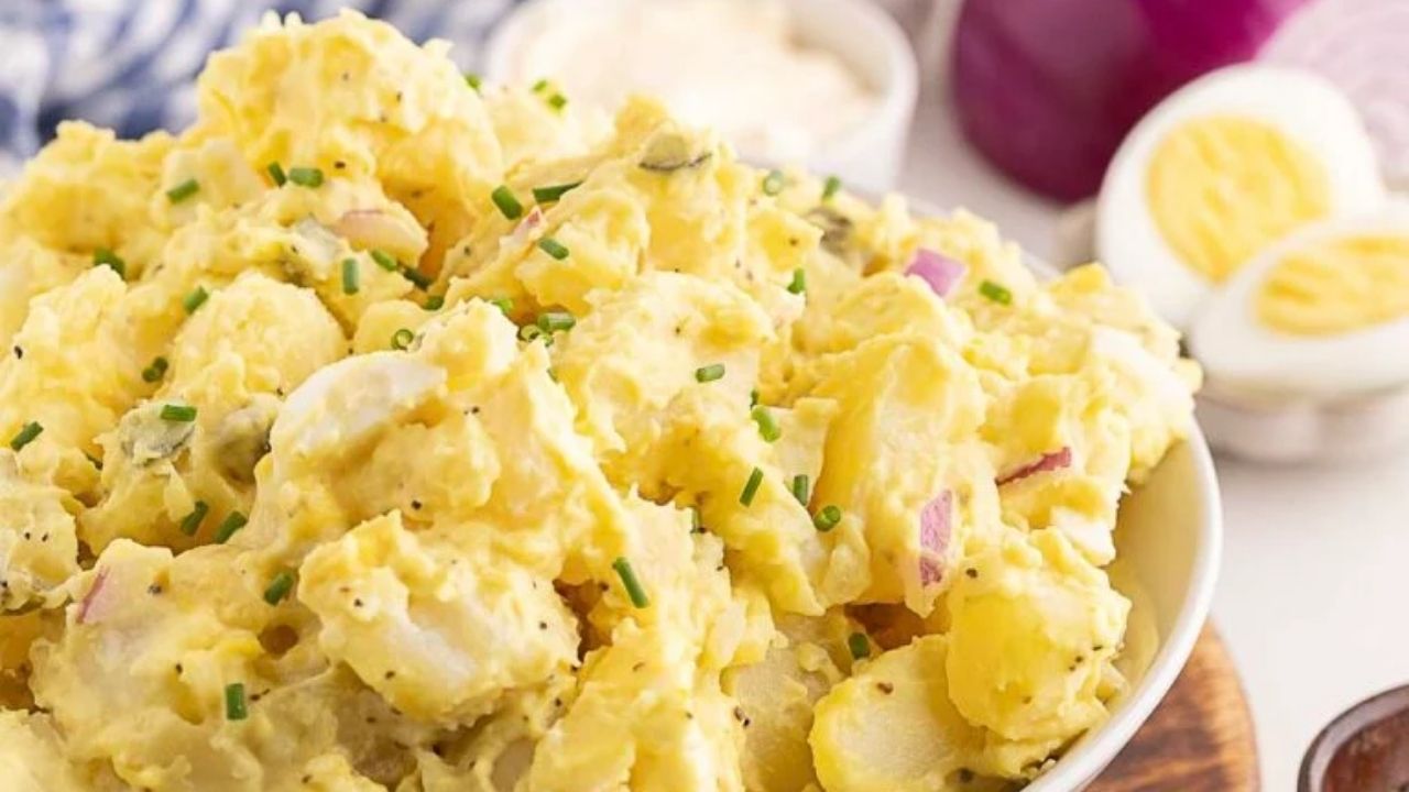 A bowl of potato salad with chopped chives and red onions, with hard-boiled eggs and a small dish of sauce in the background—perfect for serving as one of your favorite Southern summer side dishes.
