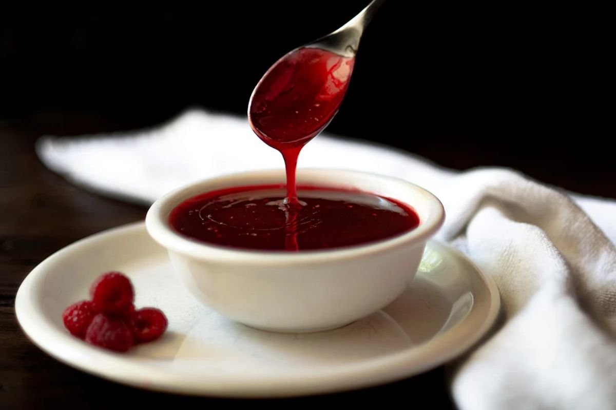A spoon drizzles red raspberry sauce into a white bowl on a plate with fresh raspberries beside it, set against a dark background.