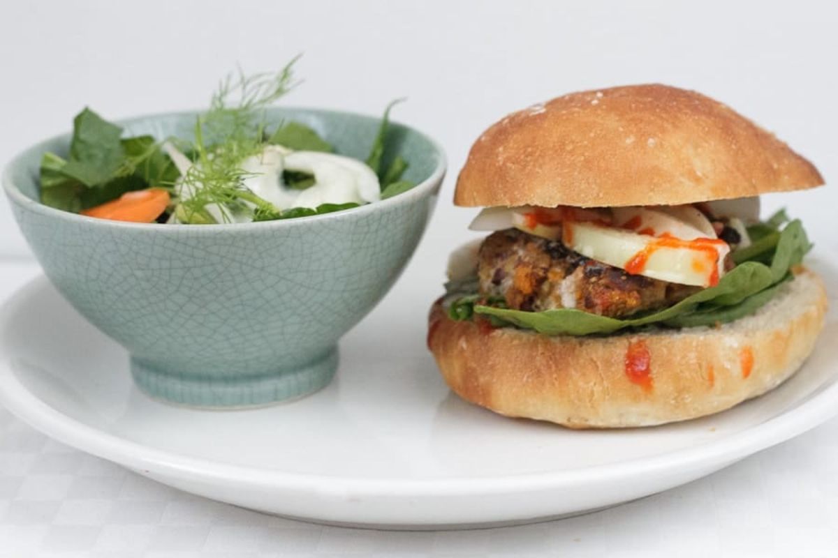 A plate with a bowl of salad containing greens and vegetables next to a sandwich with leafy greens, a patty, cheese, and sauce in a round bun.