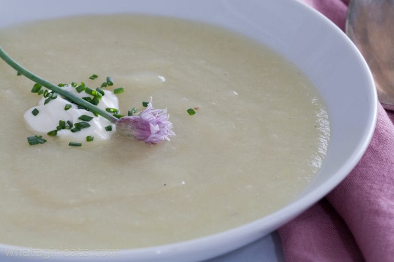 A bowl of creamy soup garnished with a dollop of cream, chopped chives, and a chive blossom, placed on a pink napkin.