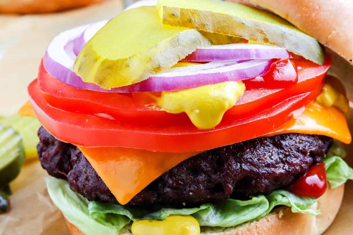 Close-up of a cheeseburger with lettuce, tomato, red onion, pickles, cheddar cheese, ketchup, and mustard in a bun.