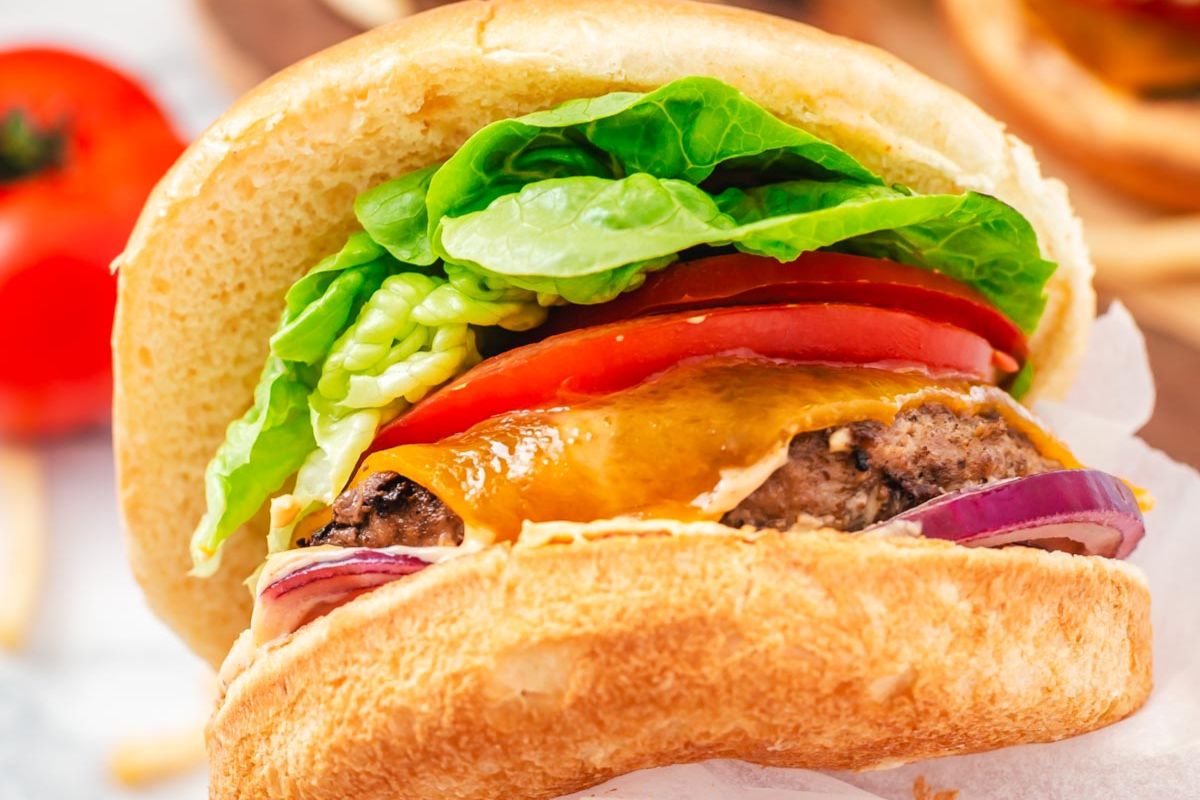 Close-up of a cheeseburger with lettuce, tomato, cheddar cheese, red onion, and a beef patty in a bun.