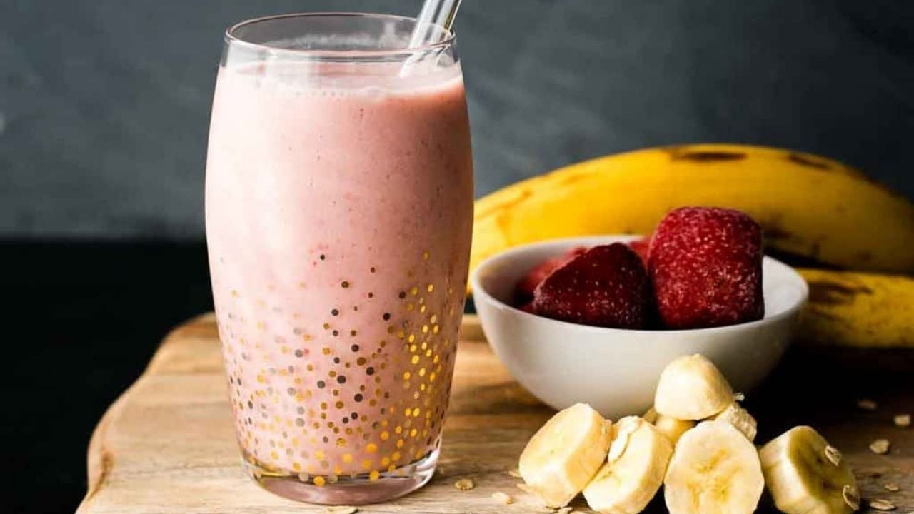 A glass of pink smoothie with a straw sits on a wooden surface, next to sliced bananas, whole bananas, and a bowl of strawberries.