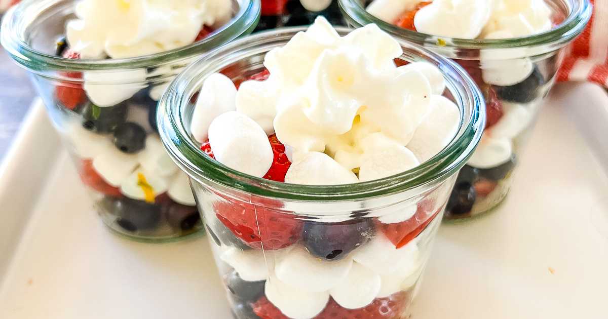 Three glass jars filled with layers of blueberries, strawberries, mini marshmallows, and topped with whipped cream, arranged on a white tray.