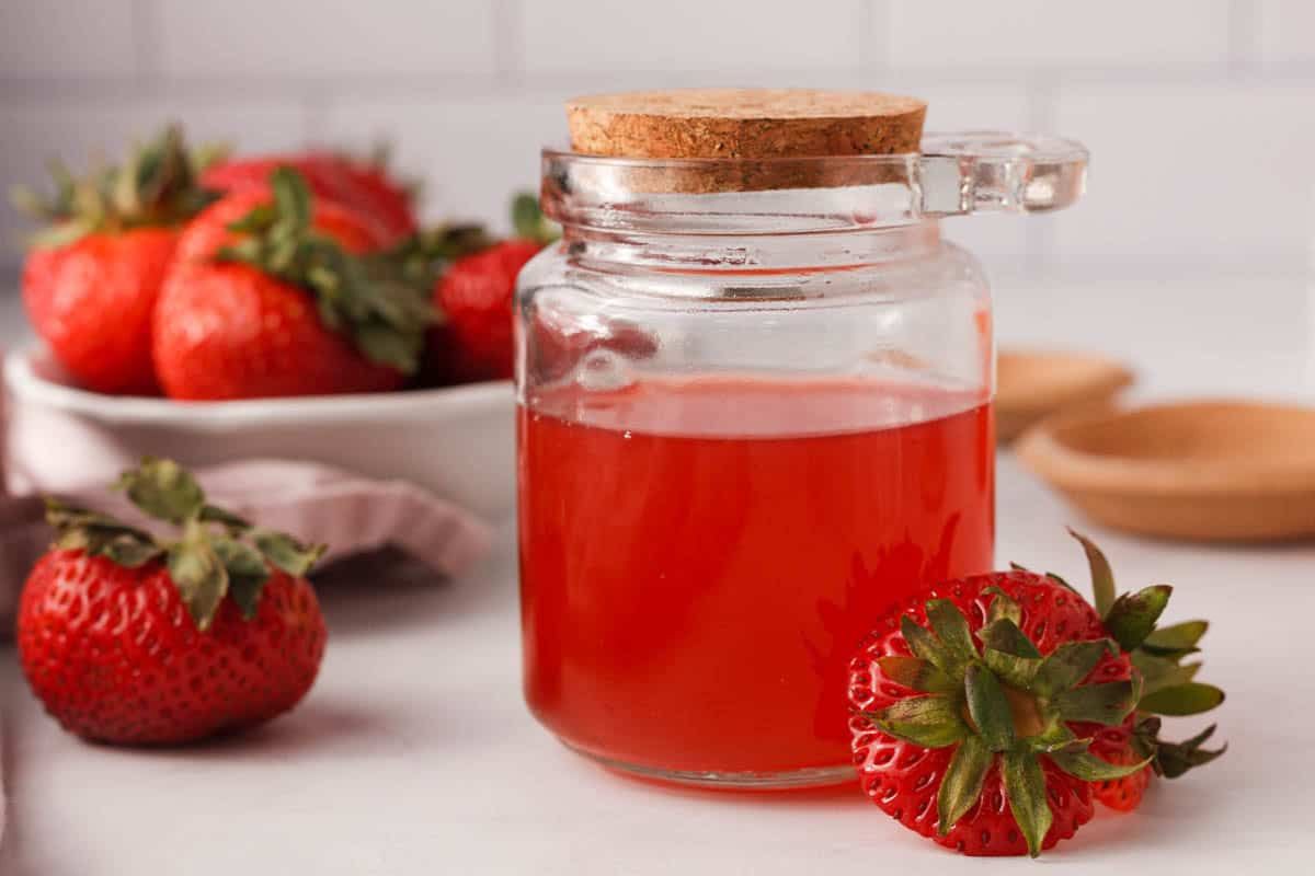 A glass jar filled with red strawberry syrup, sealed with a cork lid, sits on a counter next to fresh strawberries and a bowl of strawberries in the background.
