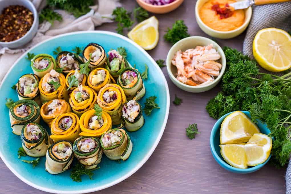 A plate of assorted vegetable rolls garnished with herbs, surrounded by bowls of dip, shredded chicken, lemon wedges, and parsley on a wooden table.