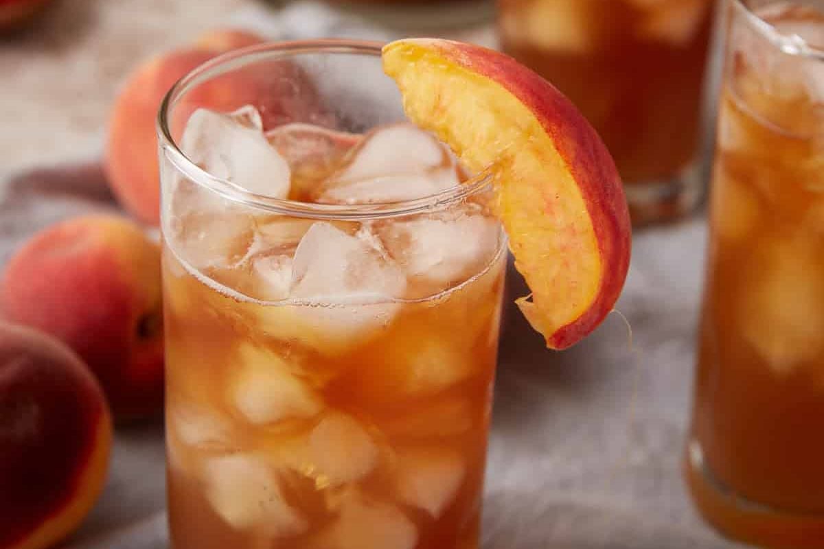 A glass of iced tea with a fresh peach slice garnish, filled with ice cubes, sits on a table next to whole peaches