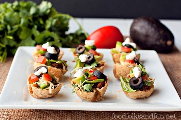 Mini taco cups filled with lettuce, diced tomatoes, olives, shredded cheese, avocado, and sour cream, arranged on a white square plate with vegetables in the background.
