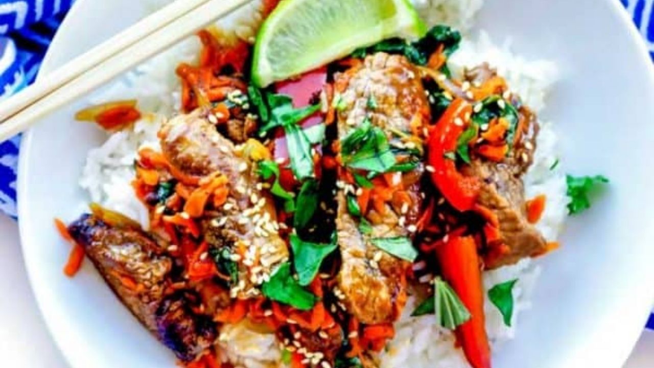 A bowl of rice topped with stir-fried beef, red bell peppers, carrots, greens, sesame seeds, and a lime wedge, with chopsticks resting on the bowl.