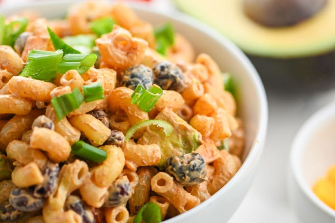 A bowl of macaroni salad with chopped green onions, raisins, and a creamy dressing, with half an avocado and a yellow bowl visible in the background.