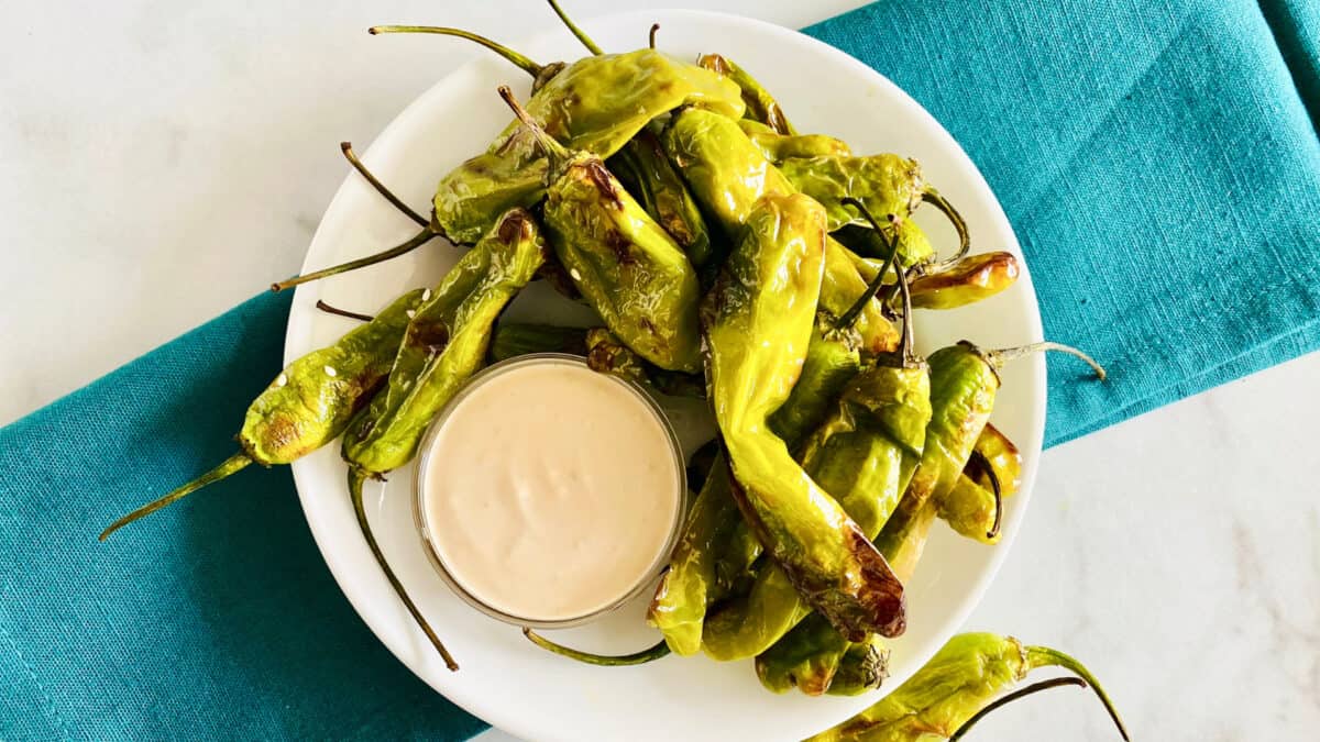 A white plate with roasted shishito peppers and a small dish of creamy dipping sauce, set on a teal napkin.