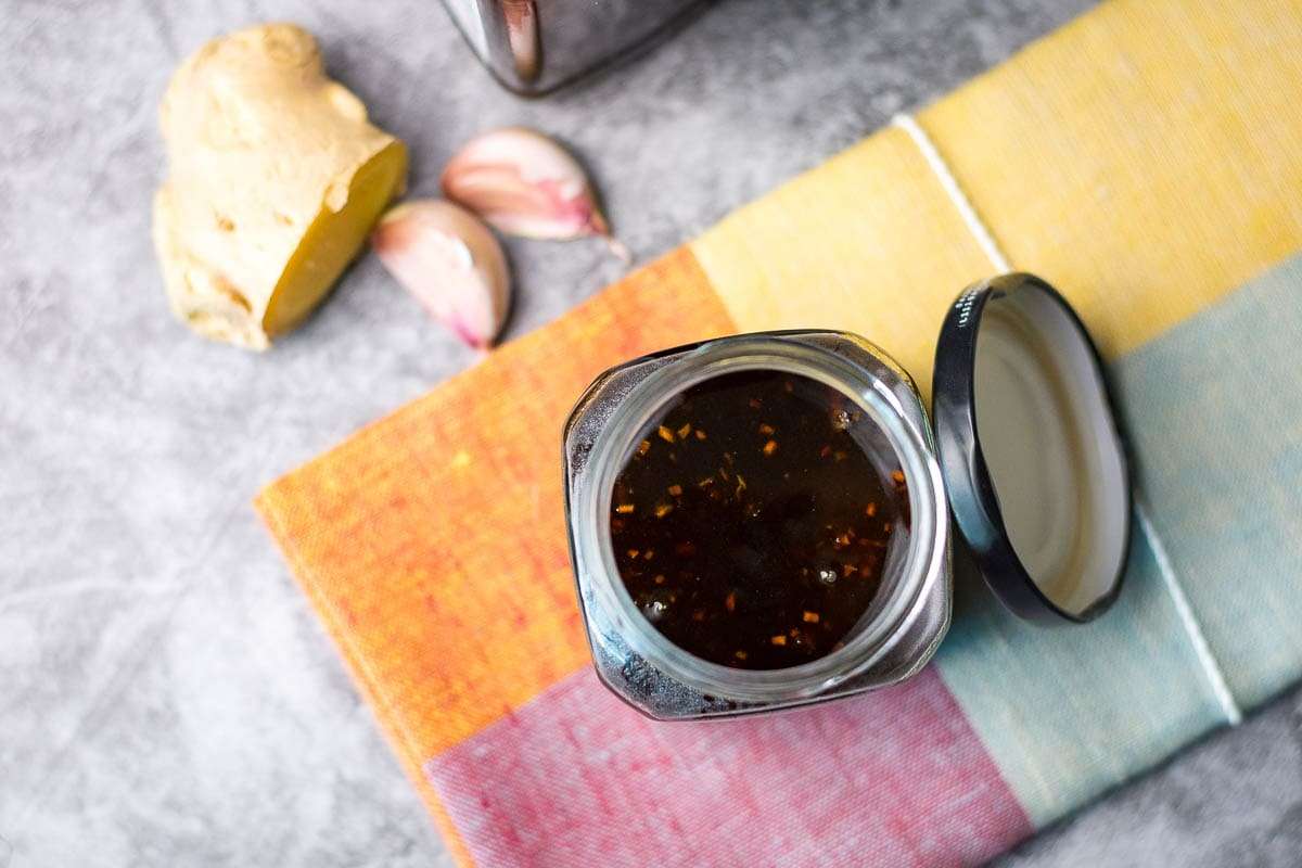 An open jar of dark sauce sits on a colorful cloth next to a piece of ginger and two garlic cloves on a gray surface.