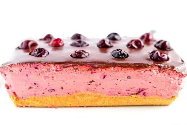 Rectangular cherry mousse cake with a yellow base, pink cherry filling, chocolate glaze, and whole cherries on top, set against a white background.