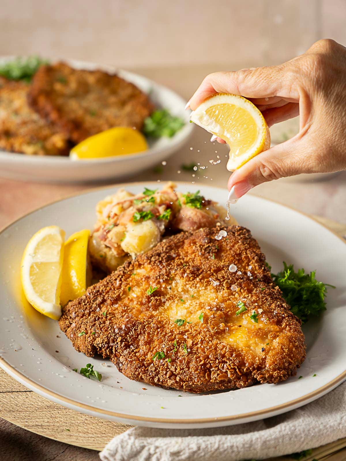 A hand squeezes a lemon wedge over a breaded, fried cutlet on a plate, garnished with parsley and served with potato salad and another lemon wedge.
