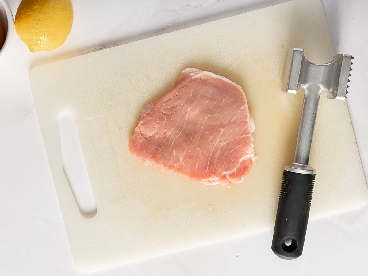 Raw pork cutlet on a white cutting board next to a meat tenderizer and part of a lemon visible in the top left corner.