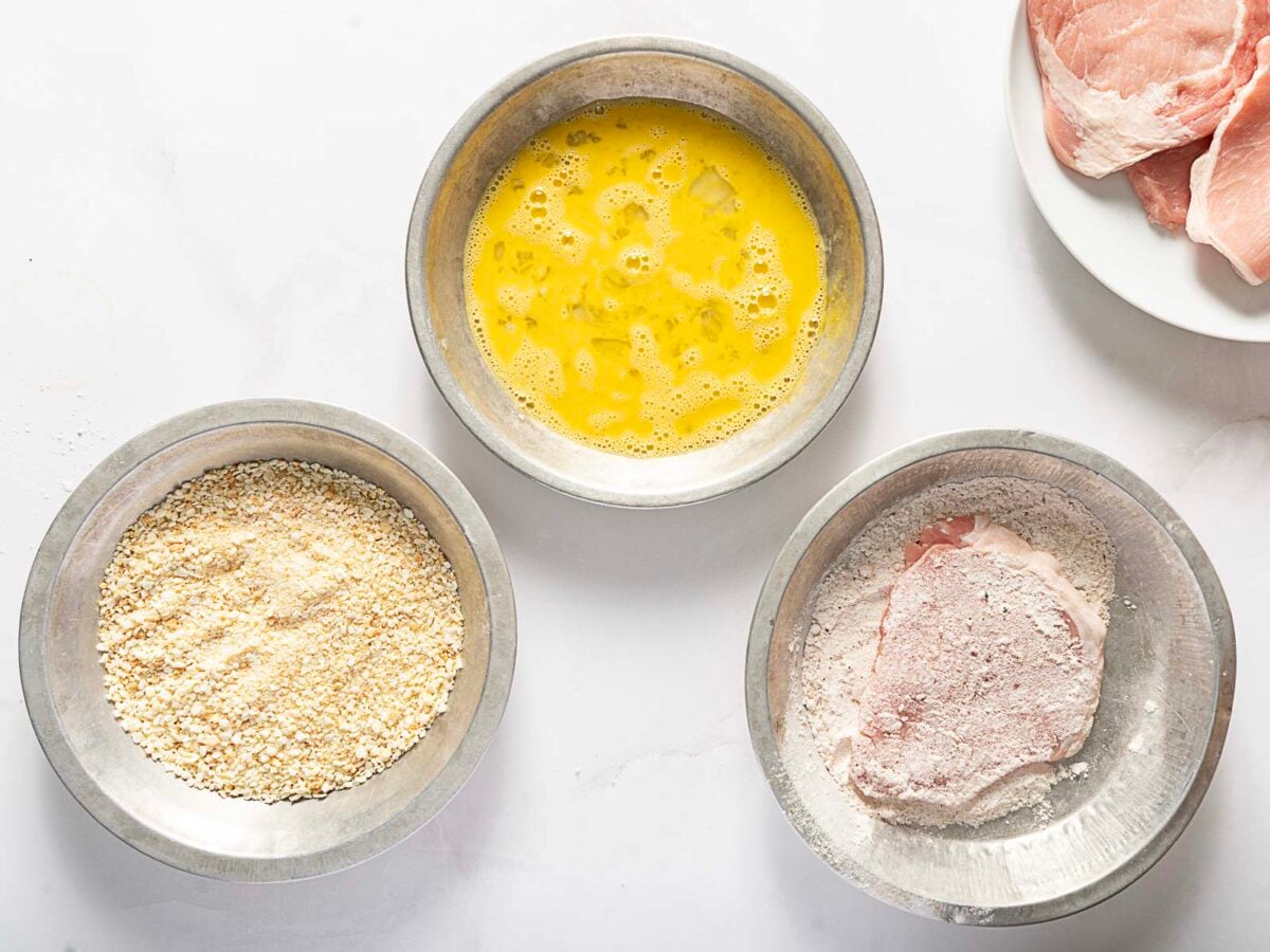 Three metal bowls with breadcrumbs, beaten eggs, and flour-coated meat; a plate with raw meat slices is partially visible on the right.
