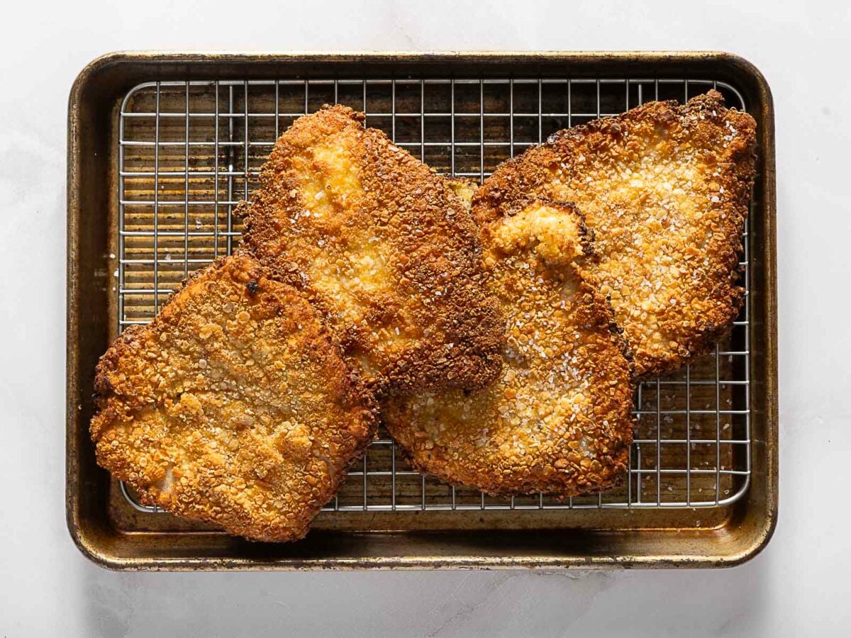 Four breaded and fried pork chops rest on a wire rack set inside a baking sheet.