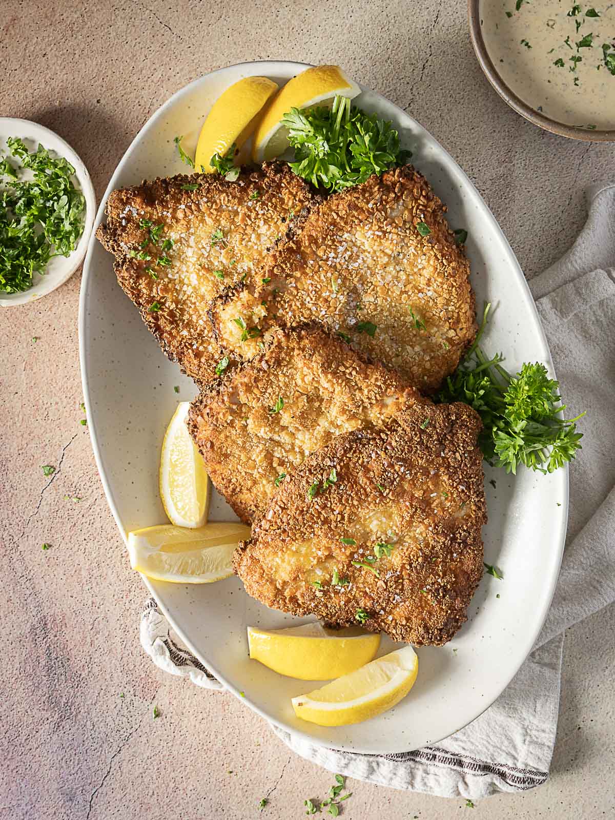 Three breaded and fried cutlets on an oval plate, garnished with parsley and lemon wedges, with a bowl of sauce and chopped herbs nearby.