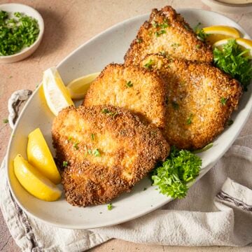 Three breaded and fried schnitzel cutlets are served on a white oval plate with lemon wedges and parsley garnish.