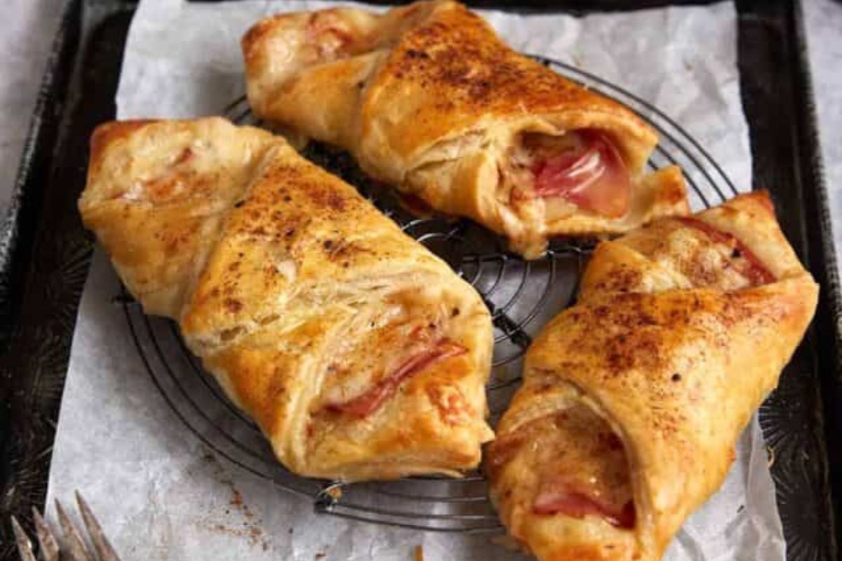 Three golden-brown puff pastry turnovers with visible ham and cheese filling, placed on a round cooling rack lined with parchment paper