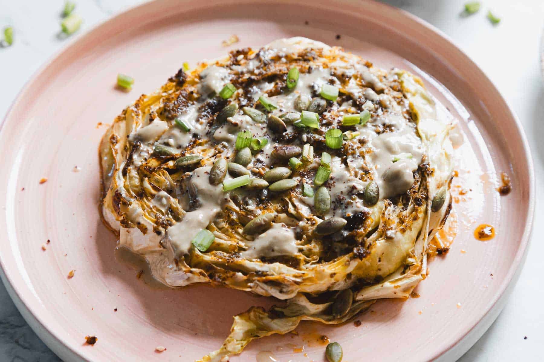 A roasted cabbage steak topped with creamy sauce, pumpkin seeds, and chopped green onions on a pink plate.