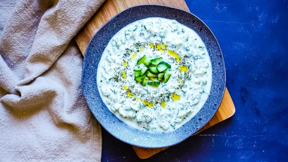 A bowl of creamy tzatziki garnished with chopped cucumber, herbs, and a drizzle of olive oil, set on a dark plate with a napkin and wooden board in the background.