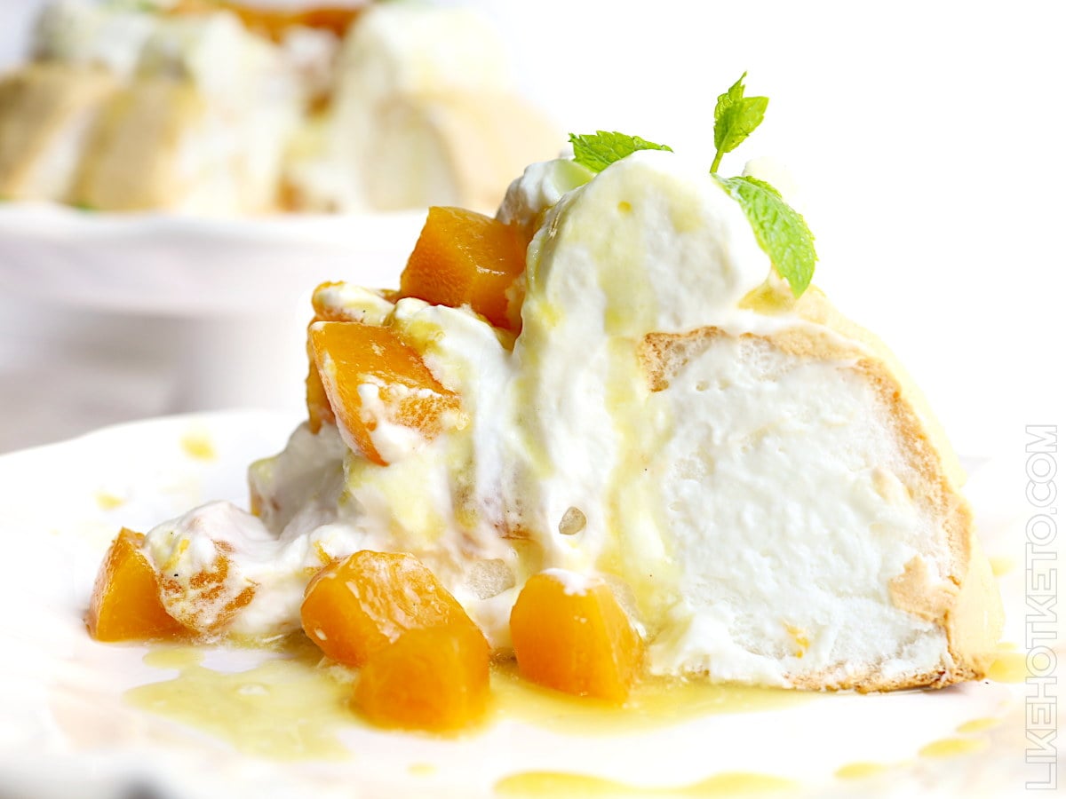 A slice of meringue dessert topped with whipped cream, diced peaches, yellow sauce, and a sprig of mint on a white plate.