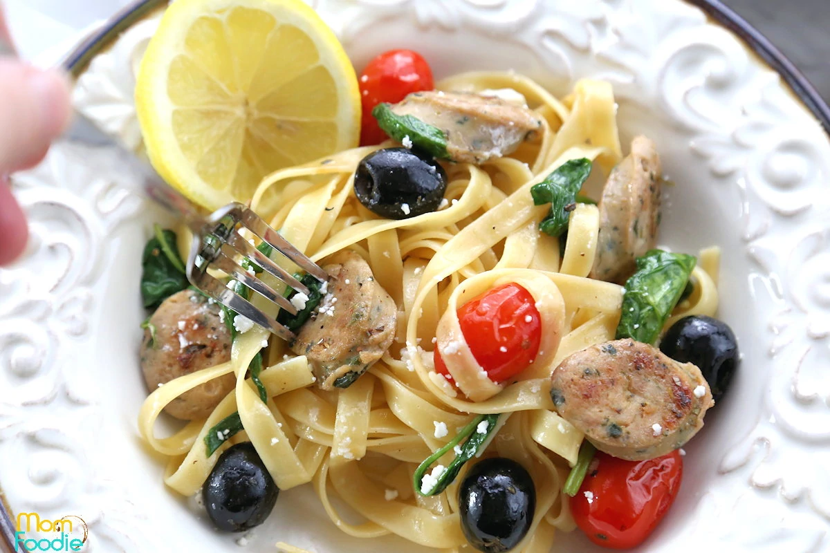 A bowl of fettuccine pasta with chicken, spinach, cherry tomatoes, black olives, feta cheese, and a lemon slice. A fork is twirling some pasta.