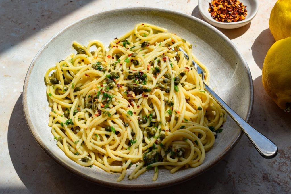 A bowl of spaghetti with capers, herbs, red pepper flakes, and olive oil, served with a fork. Lemon and a small dish of red pepper flakes are nearby.
