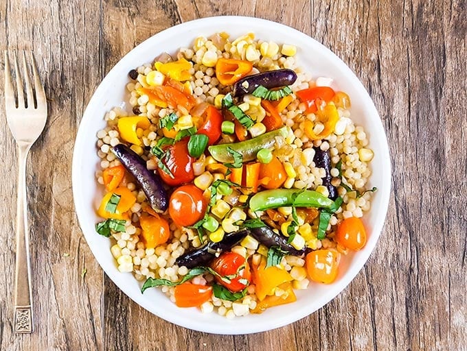 A plate of Israeli couscous salad with cherry tomatoes, bell peppers, eggplant, corn, and fresh herbs on a wooden table, fork nearby.