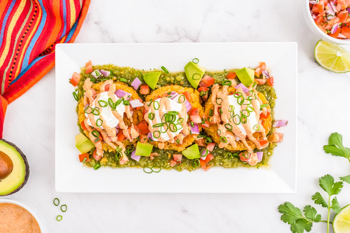 Three tostadas topped with diced tomatoes, red onion, avocado, sour cream, and green onions are arranged on a rectangular plate over a green sauce. Other ingredients are around the plate.
