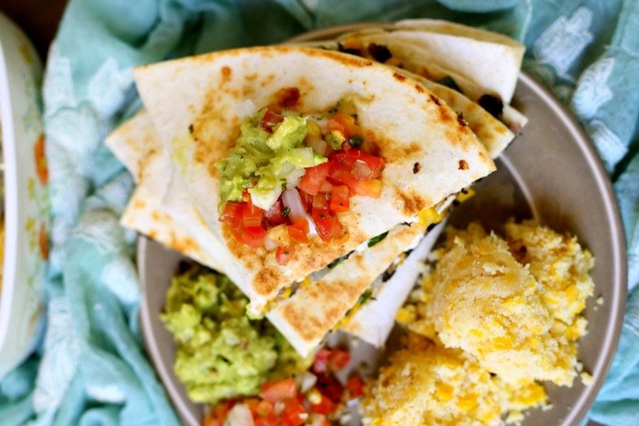 A plate with quesadilla slices topped with guacamole and salsa, served with extra guacamole, salsa, and two scoops of cornbread.