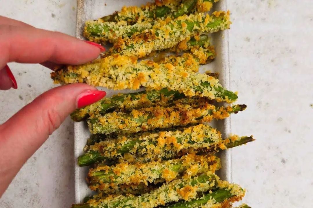 A hand with red nail polish picks up a breaded and baked green bean from a plate of similar green bean fries.