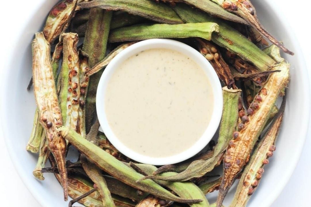 A bowl of creamy dipping sauce is surrounded by roasted okra halves on a white plate.