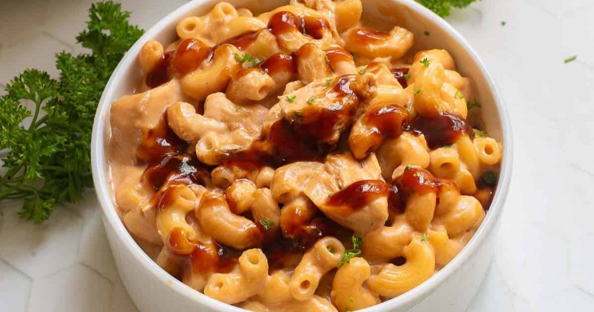 Bowl of macaroni and cheese mixed with chunks of chicken and topped with a drizzle of barbecue sauce, garnished with parsley.