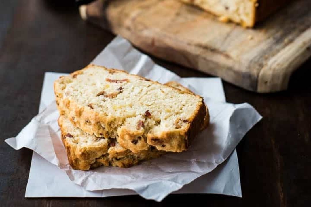 Two slices of bread with visible bits of bacon rest on parchment paper, with a wooden cutting board holding more bread in the background.