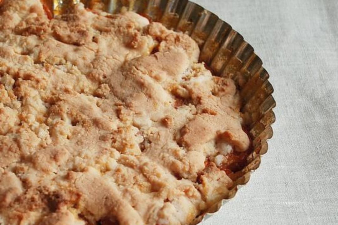 A golden-brown crumbly pie or tart sits in a fluted metal baking tin on a light-colored fabric surface.
