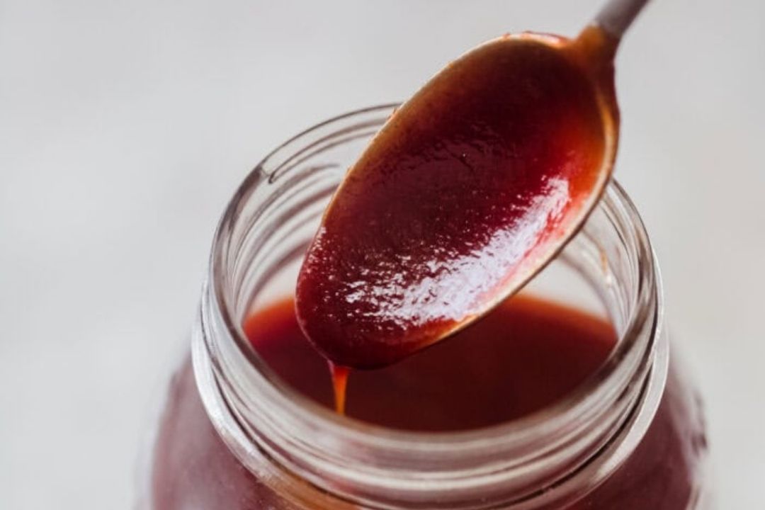 A spoon with reddish-brown sauce held above an open glass jar filled with the same sauce.